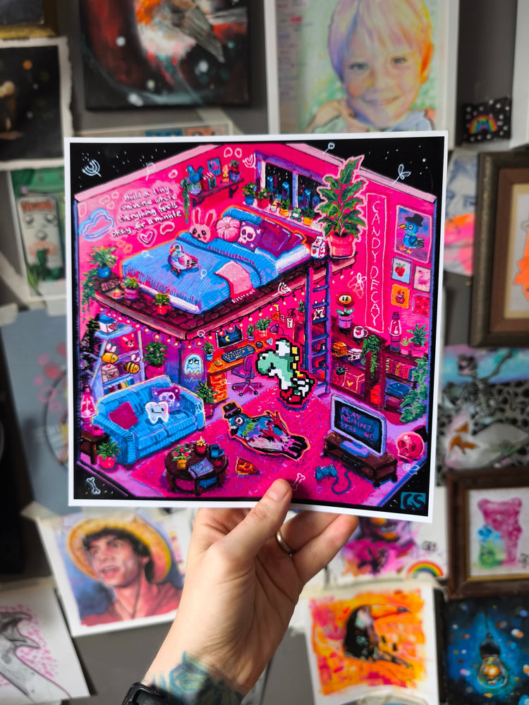 Hand holding a colorful art print titled “In My Candy Decay Apartment,” depicting a surreal neon pink two-level apartment filled with plants, pigeons, skulls, retro gaming details, a pixel-art unicorn, bright blue furniture and playful objects.