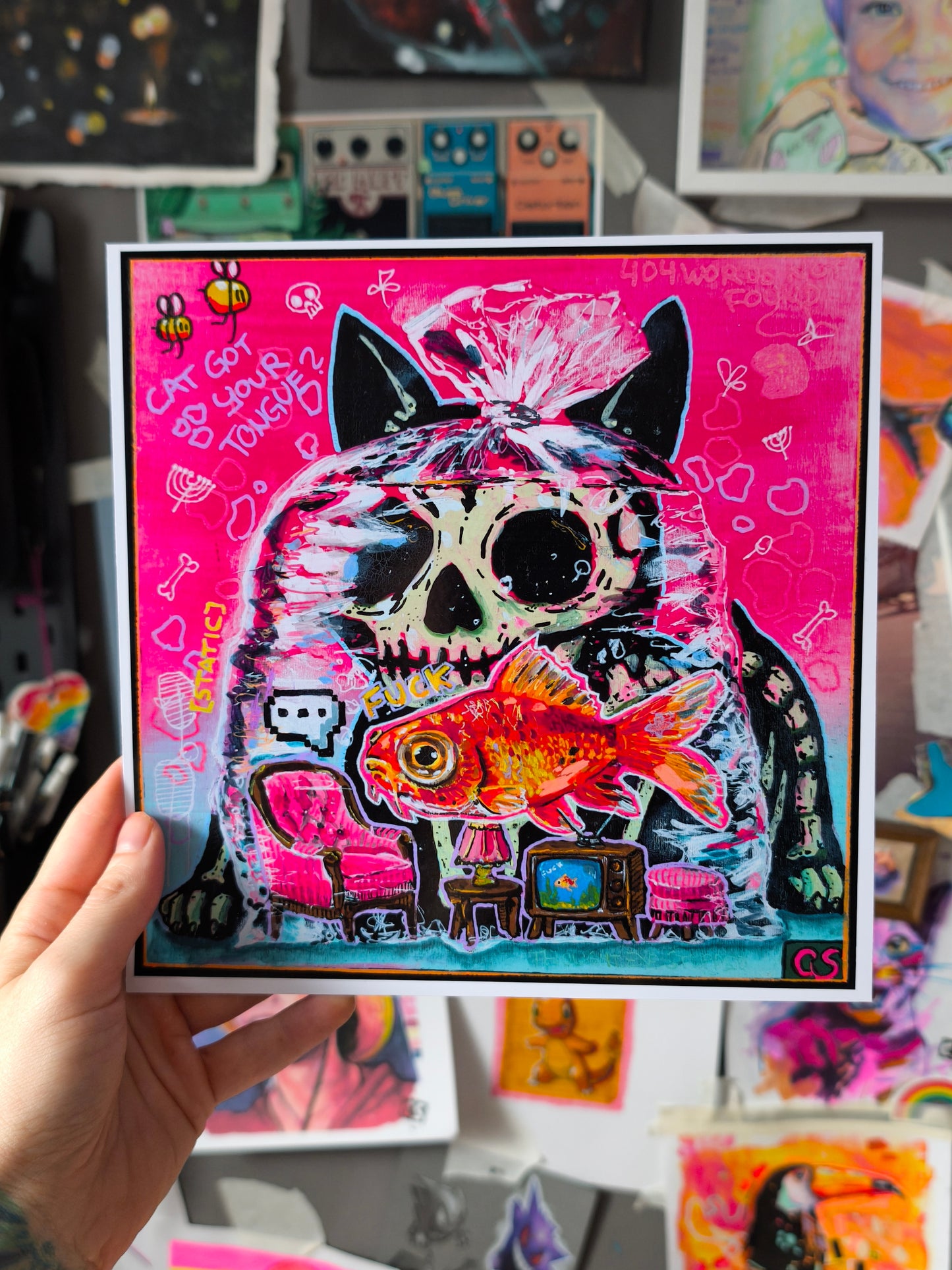 Colorful pop-surreal artwork titled Cat Got Your Tongue? featuring a large black cat with a skull-like face staring over a goldfish trapped inside a transparent plastic bag. Tiny furniture, including a pink armchair, lamp and vintage television, sits inside the bag beneath the fish. The background is neon pink and turquoise with handwritten doodles, bones, bees and playful symbols.