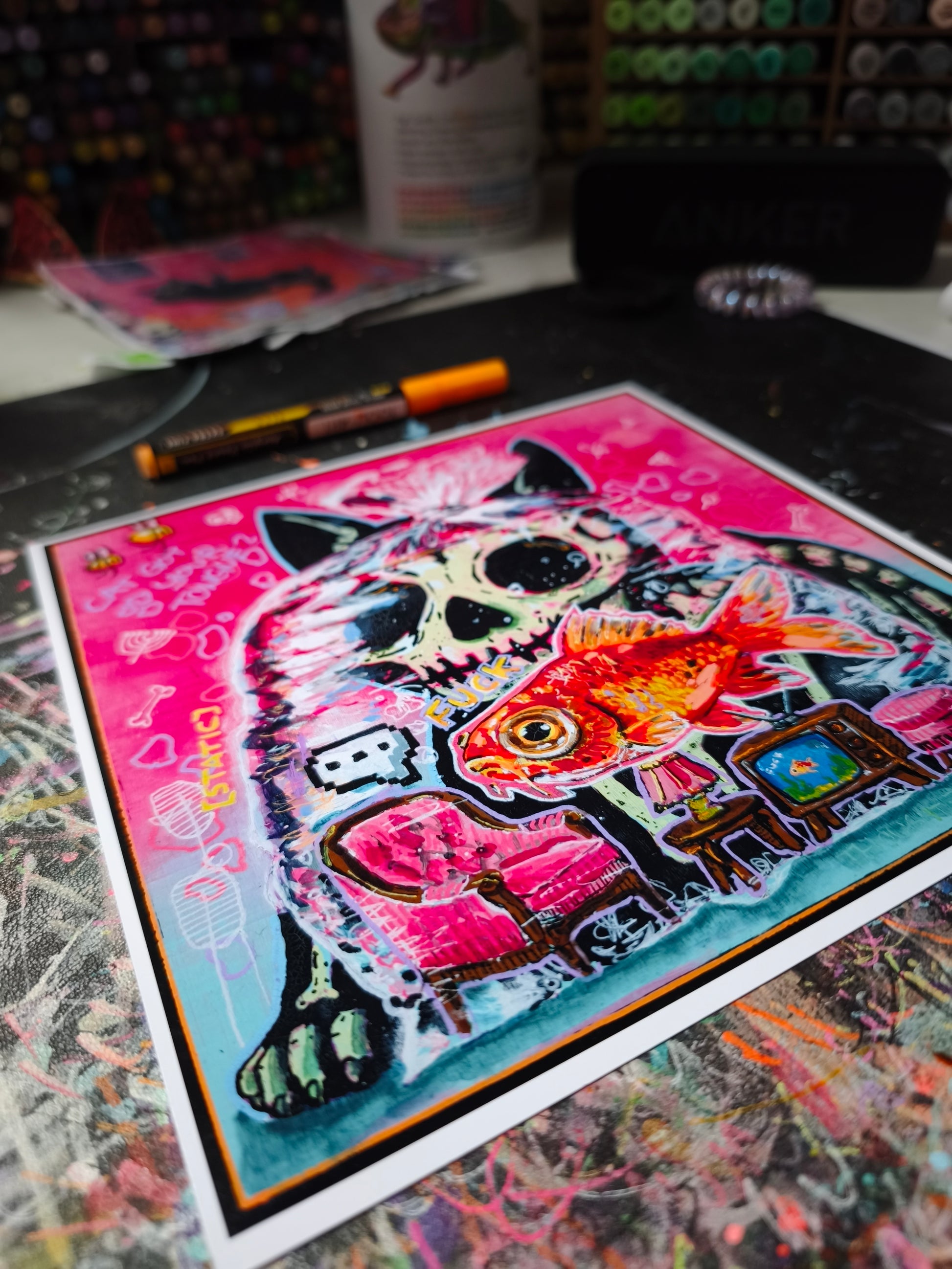 Colorful pop-surreal artwork titled Cat Got Your Tongue? featuring a large black cat with a skull-like face staring over a goldfish trapped inside a transparent plastic bag. Tiny furniture, including a pink armchair, lamp and vintage television, sits inside the bag beneath the fish. The background is neon pink and turquoise with handwritten doodles, bones, bees and playful symbols.