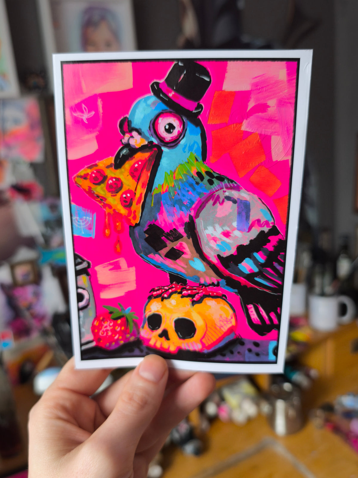 Pizza-Taube als Postkarte von Paintranger: eine bunte Taube mit schwarzem Zylinder und einem Stück Pizza im Schnabel, die vor pinkem Hintergrund auf einem Totenkopf steht